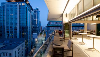 A rooftop terrace overlooking a city street with modern buildings, featuring outdoor seating and a view of the skyline at dusk.