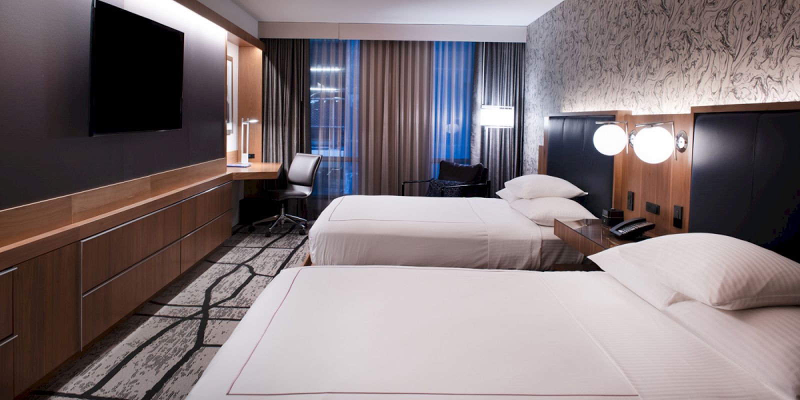 The image shows a modern hotel room with two beds, a large TV, desk, chair, and lamps. The decor is stylish and contemporary.