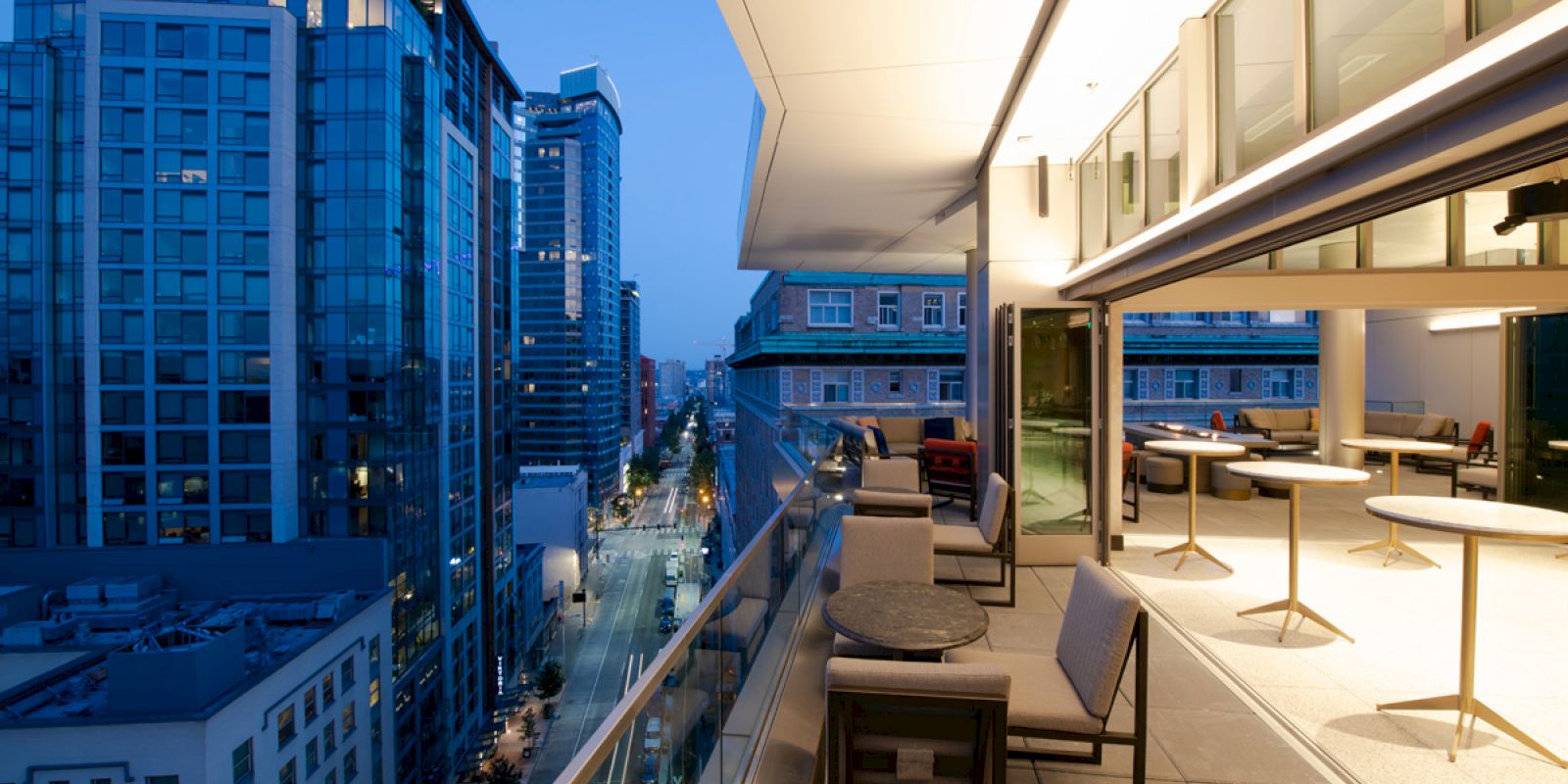 A modern cityscape with tall buildings at dusk, featuring a lit balcony with tables and chairs overlooking the street.