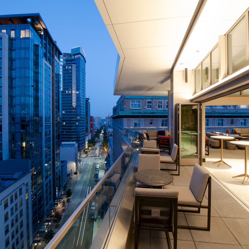 A modern balcony overlooks a cityscape at dusk, with buildings and a street below. The area features tables and seating arrangements.