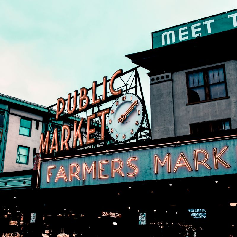 The image shows a famous marketplace with neon signs reading "Public Market" and "Farmers Market," under a clock, against an urban backdrop.