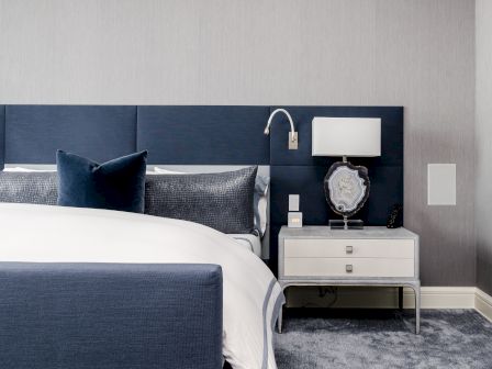 A modern bedroom features a blue upholstered bed, grey pillows, a nightstand with a lamp and decorative item, on a carpeted floor.