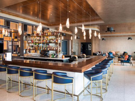 The image shows a modern bar with a wooden counter, blue bar stools, hanging lights, and shelves filled with bottles in a spacious setting.
