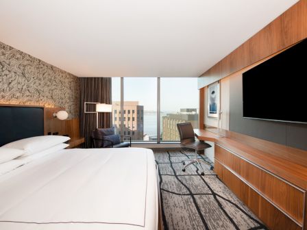 A modern hotel room featuring a large bed, flat-screen TV, desk, chair, and a window with a city view.