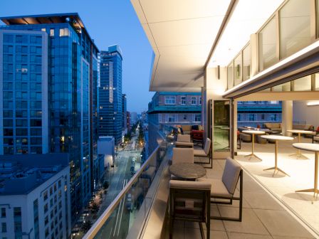 A modern cityscape view from a balcony at dusk, featuring outdoor seating and buildings with glass facades.