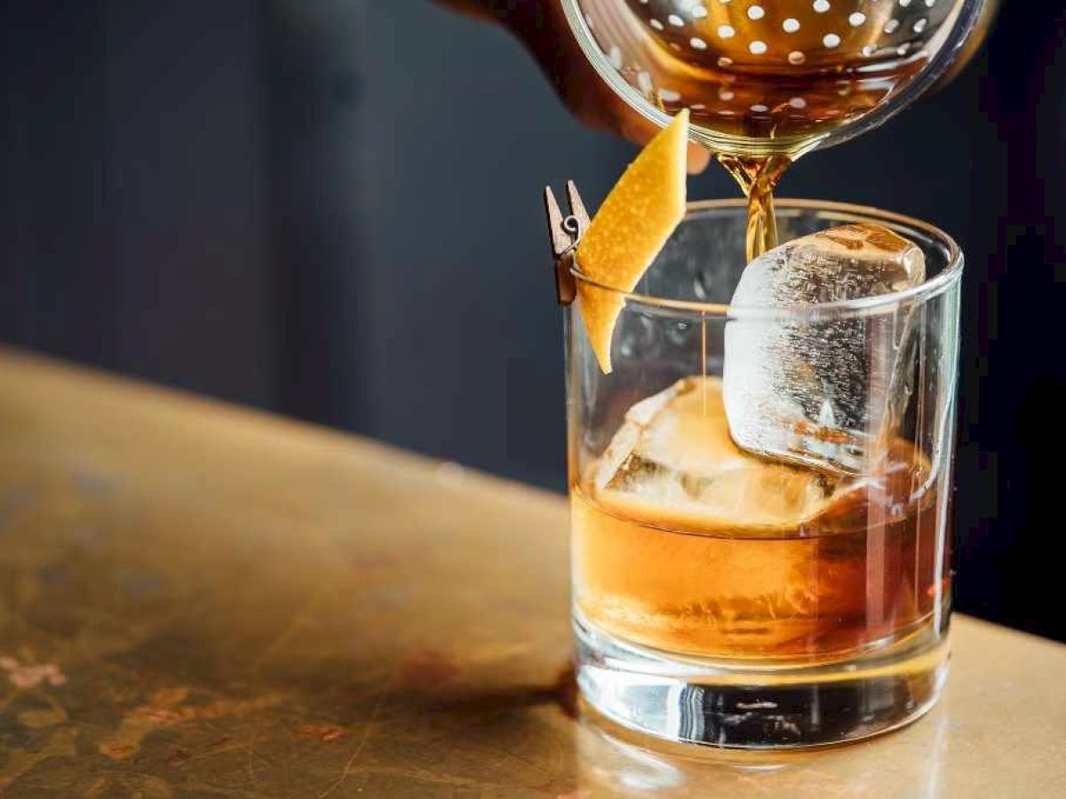 A cocktail being poured into a glass with ice and garnished with a twist of citrus peel sits on a polished surface.