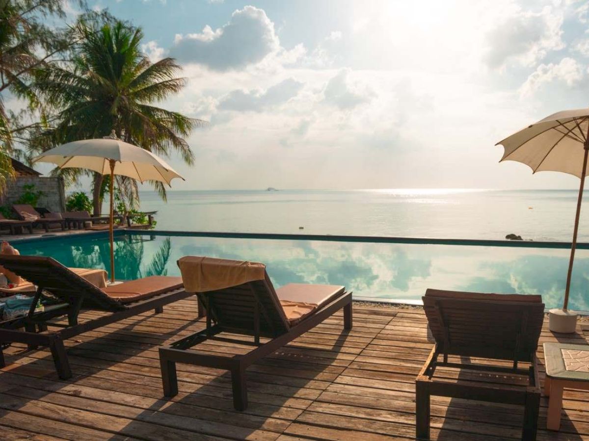 A relaxing poolside scene with lounge chairs, umbrellas, palm trees, and a view of the ocean under a partly cloudy sky.