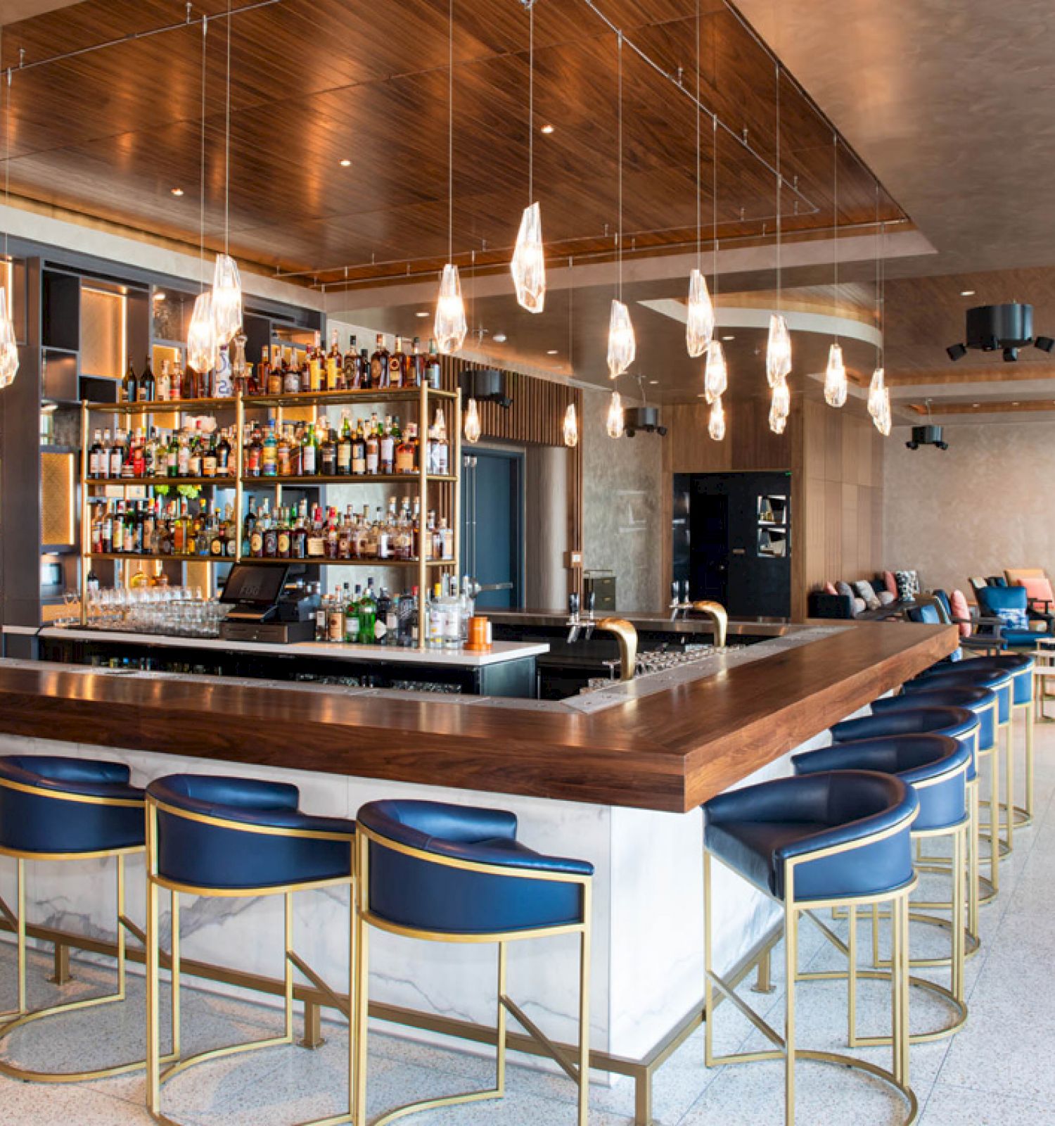 A modern bar with a wooden counter, blue cushioned stools, hanging lights, and a fully stocked back bar; lounge seating in the background.
