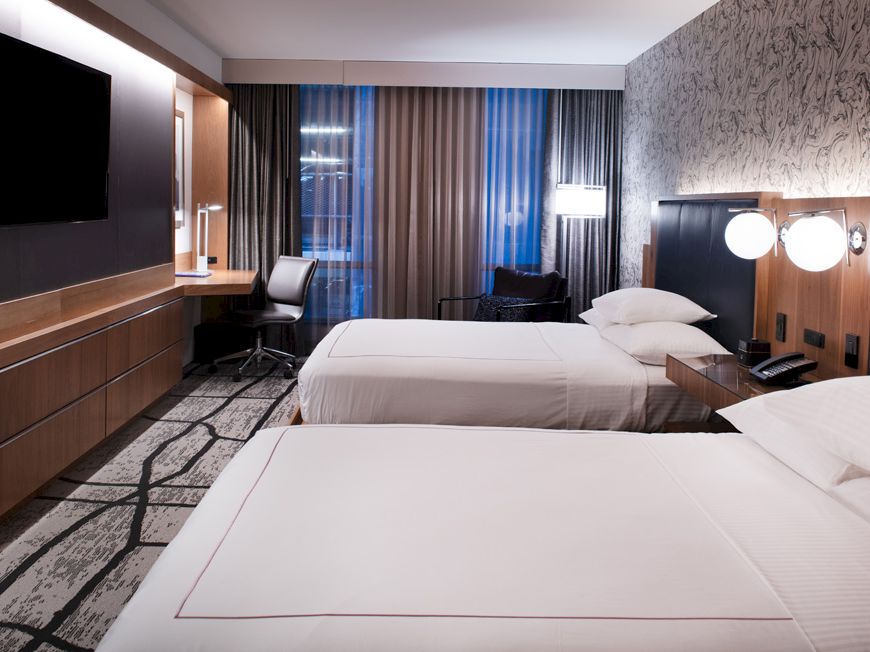 The image shows a modern hotel room with two beds, a TV, a desk with a chair, lamps, and patterned walls and carpet.