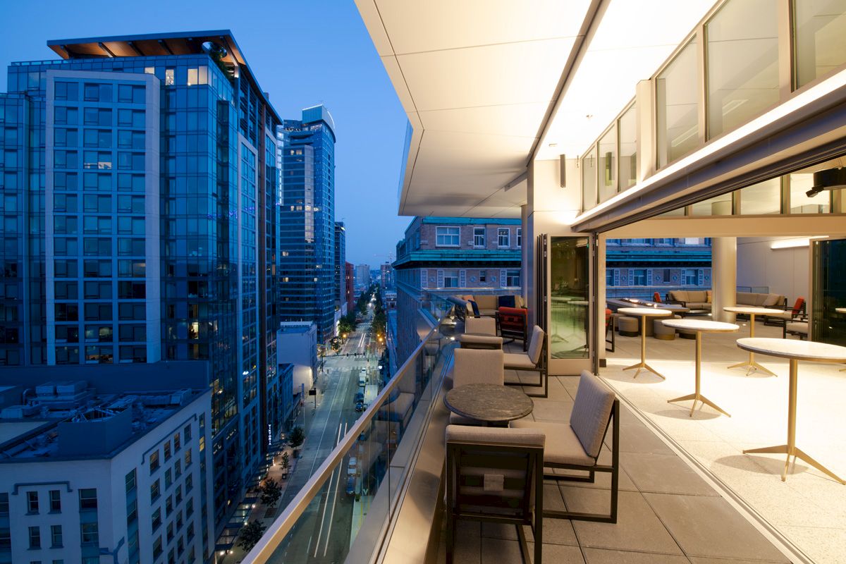 A modern rooftop patio with tables and chairs overlooks a cityscape of tall buildings at twilight, creating a sleek urban atmosphere.