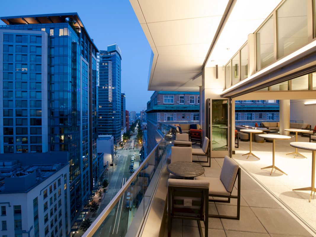 A modern rooftop patio with tables and chairs overlooks a cityscape of tall buildings at twilight, creating a sleek urban atmosphere.