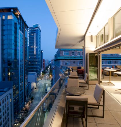 A modern cityscape with tall buildings at twilight, viewed from a stylish balcony with seating and tables, overlooking a lively street.
