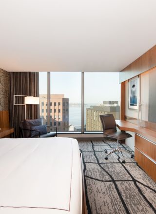 A modern hotel room with a bed, large TV, desk, and window view of buildings and water. The decor is sleek with wood and patterned walls.