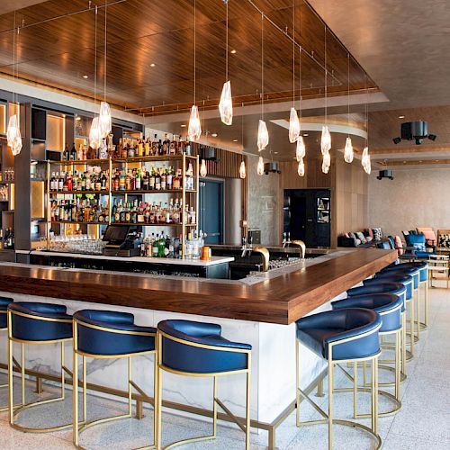 A modern bar with elegant stools, hanging lights, and a well-stocked shelf. Cozy seating area visible in the background completes the setting.