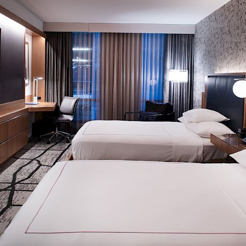 A hotel room with two beds, a TV, a desk, lamps, and modern decor.