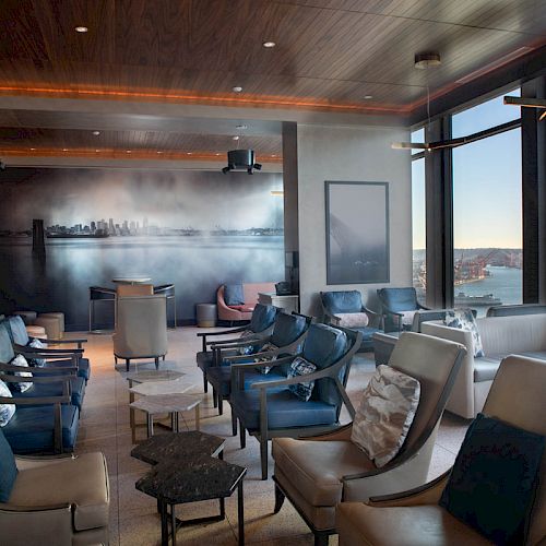 A modern lounge with plush seating, large windows offering a waterfront view, and ambient lighting. A cityscape mural adorns one wall.