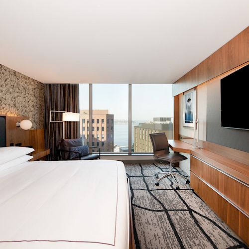 A modern hotel room with a large bed, TV, desk, and a window view of buildings and water.