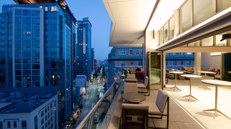 A modern rooftop terrace with seating overlooks a cityscape of high-rise buildings at dusk, featuring warm lighting and a stylish interior.