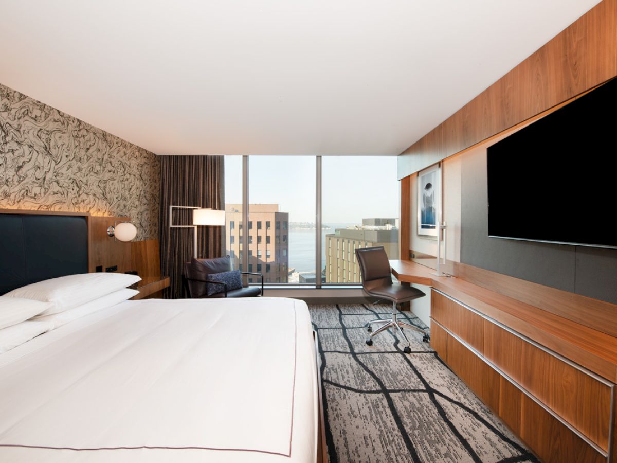 A modern hotel room with a large bed, desk, and chair facing a flat-screen TV. A window offers a cityscape view.