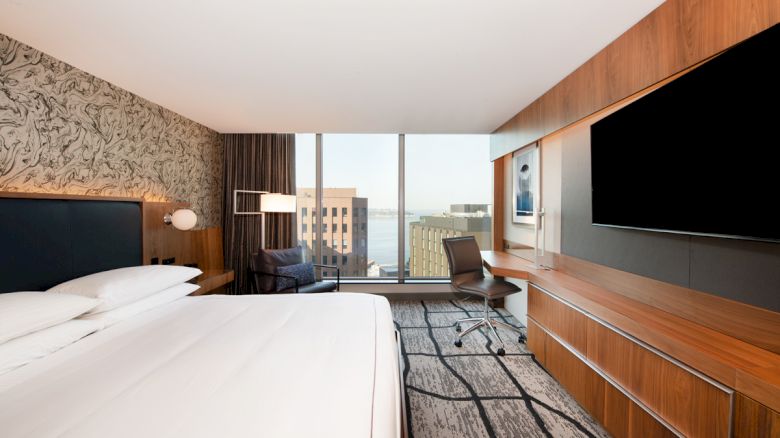 A modern hotel room with a large bed, desk, and chair facing a flat-screen TV. A window offers a cityscape view.