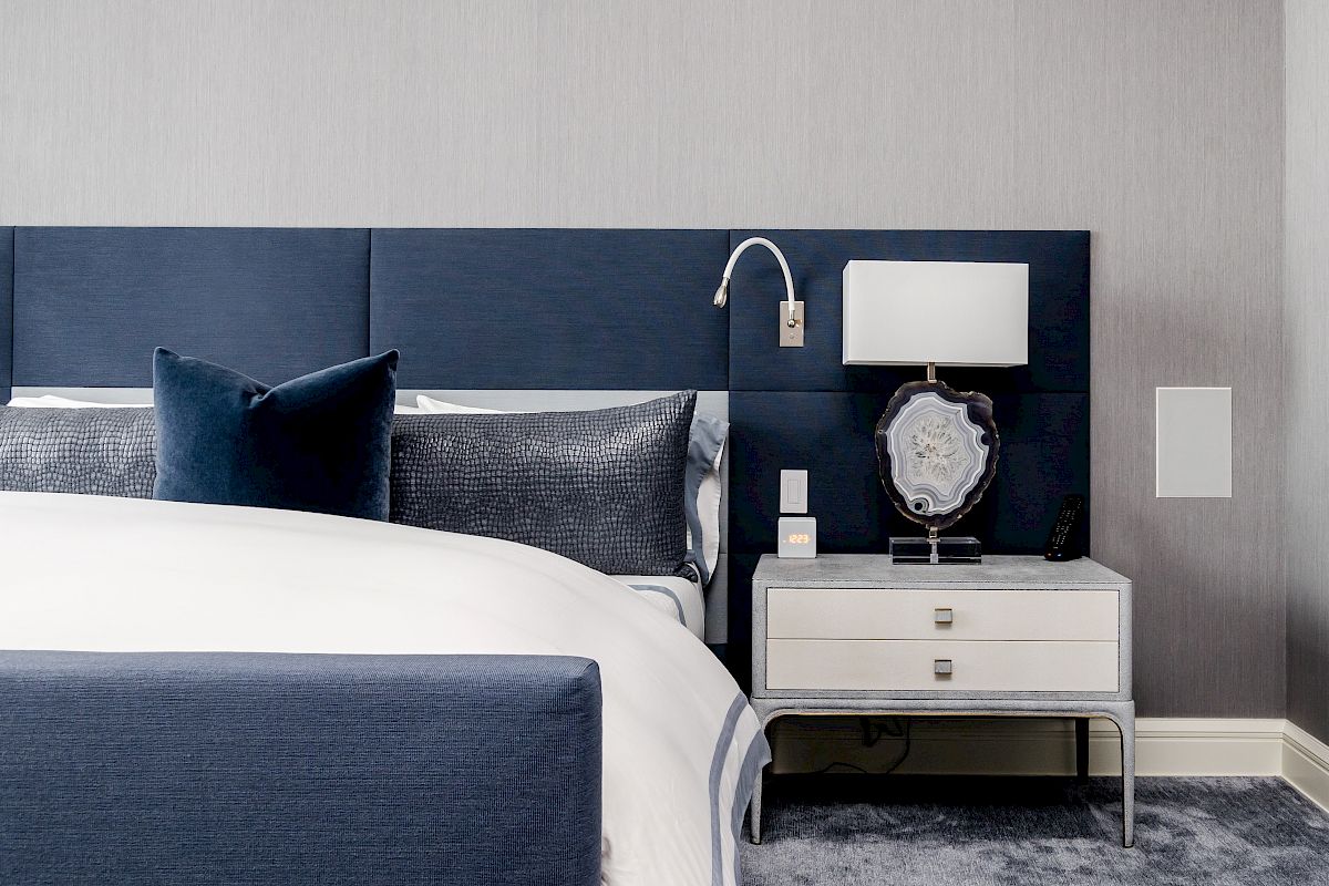 A modern bedroom with a blue upholstered headboard, white bedding, and a side table holding a lamp and decorative object.