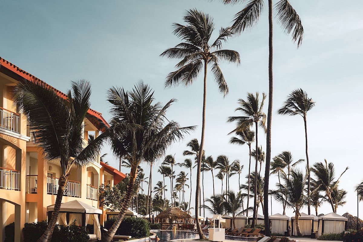 A tropical resort scene with tall palm trees, a reflective swimming pool, lounge chairs, and a building with balconies under a clear sky.
