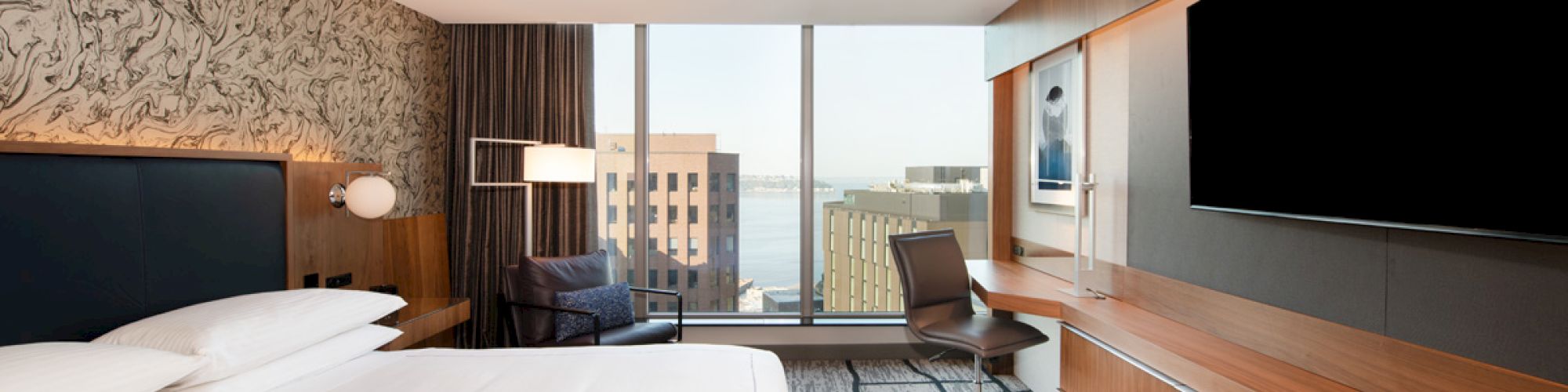 A modern hotel room features a large bed, a wall-mounted TV, a desk with a chair, and a view of buildings through a large window.